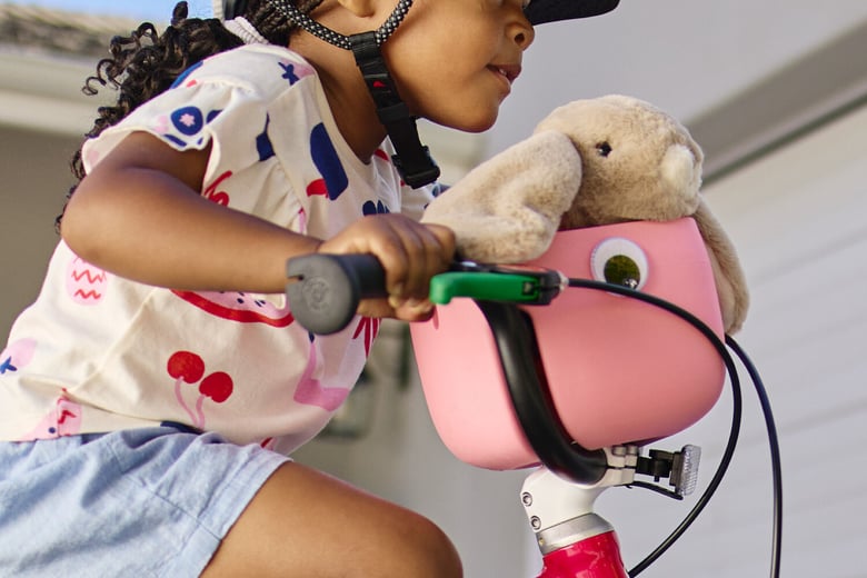A young girl wearing a woom READY helmet rides a woom red GO bike with attached marshmallow pink POP basket holding a stuffed rabbit toy.