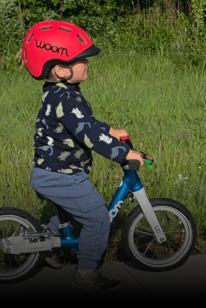 Barn med rød READY cykelhjelm på hovedet ruller afsted på en blå løbecykel fra woom, i baggrunden ses græs.
