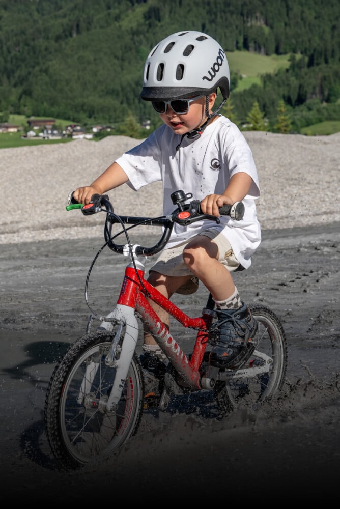 Barn iført grå READY hjelm og sommertøj kører gennem vand på en rød woom børnecykel.