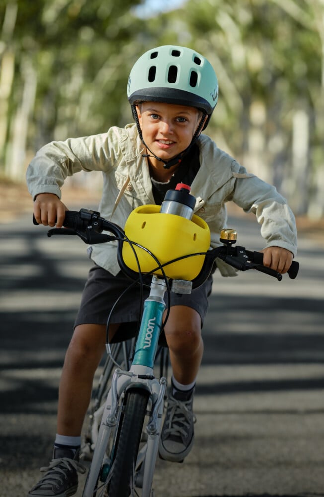 A boy wearing a woom READY helmet, riding a metallic turquoise woom GO bike with vibrant yellow POP basket holding a GLUG water bottle while riding on a road.