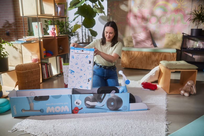Eine Frau kniet hinter der Verpackung des woom WOW Laufrades und zieht einen Teil der Innenverpackung nach oben aus der Kartonage.