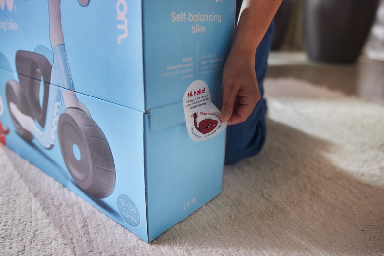 Blaue Verpackung des woom WOW Laufrads. Eine Hand öffnet die Kartonage an der vorgesehenen Stelle.