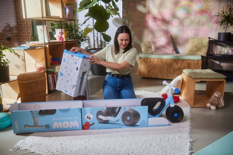 Eine Frau kniet hinter der Verpackung des woom WOW Laufrades, das Rad rollt aus der Verpackung.