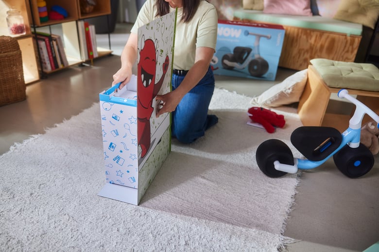 Eine Frau kniet in ihrem Wohnzimmer auf dem Teppichboden. Sie stellt die blaue Box der woom WOW Laufrad-Verpackung auf die gefaltete Fläche des Buddy-Aufstellers.