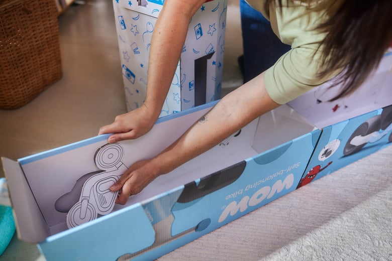 Man sieht das Innenleben der Verpackung des woom WOW Laufrades. Zwei Hände einer Frau sind im Bild, welche Papp-Aufsteller anhand einer perforierten Linie aus der Kartonage heraustrennt.