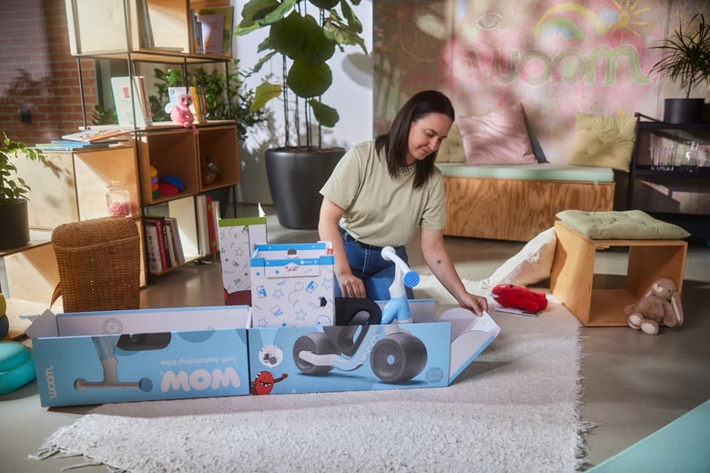 Eine Frau kniet hinter der Verpackung des woom WOW Laufrades und klappt die vordere Lasche der Kartonage nach unten, damit das Laufrad aus der Verpackung rollen kann.