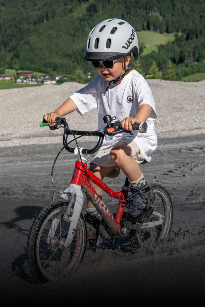 A child wearing a gray READY helmet and summer clothing is riding a red woom children's bike through water.