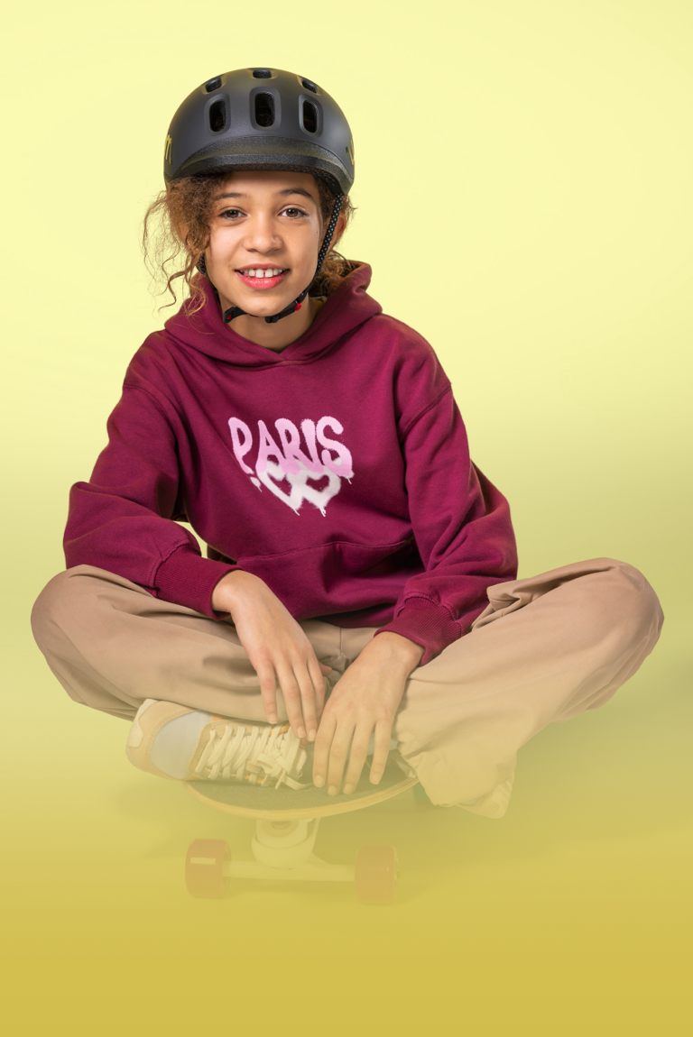 Mädchen mit schwarzem READY Kinderhelm von woom auf dem Kopf sitzt mit gekreuzten Beinen auf einem Skateboard.