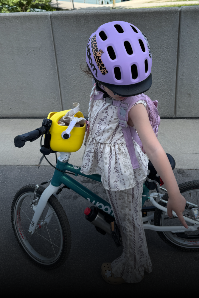 A child wearing a purple READY bike helmet is standing over a green woom bike with a yellow POP bike basket.