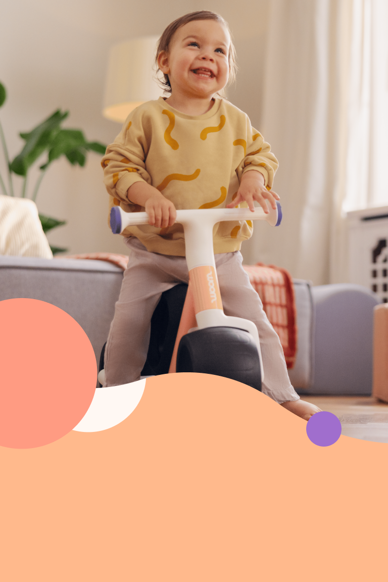 A toddler sits barefoot on the woom WOW balance bike in the living room, holding onto the handlebars and laughing happily.
