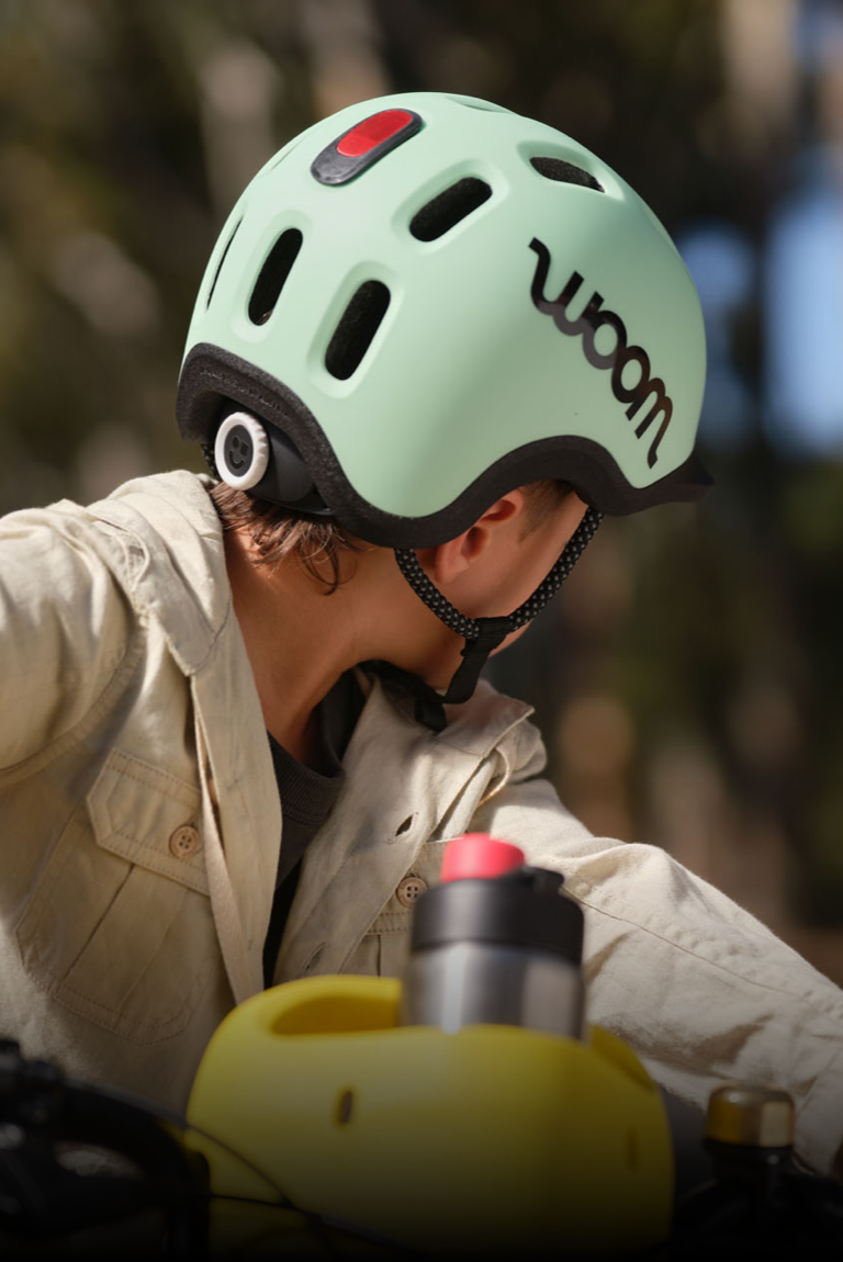 A boy in a mint green READY Kids' Helmet by woom with a clipped-in helmet light turns his head away from the camera
