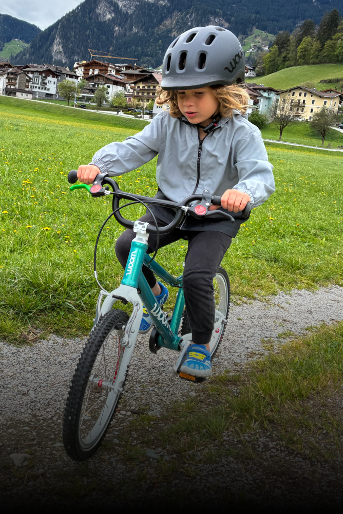 Kind mit schwarzem READY Helm fährt mit einem grünen woom GO Fahrrad auf einem Schottertrail durch eine Wiese in den Bergen.