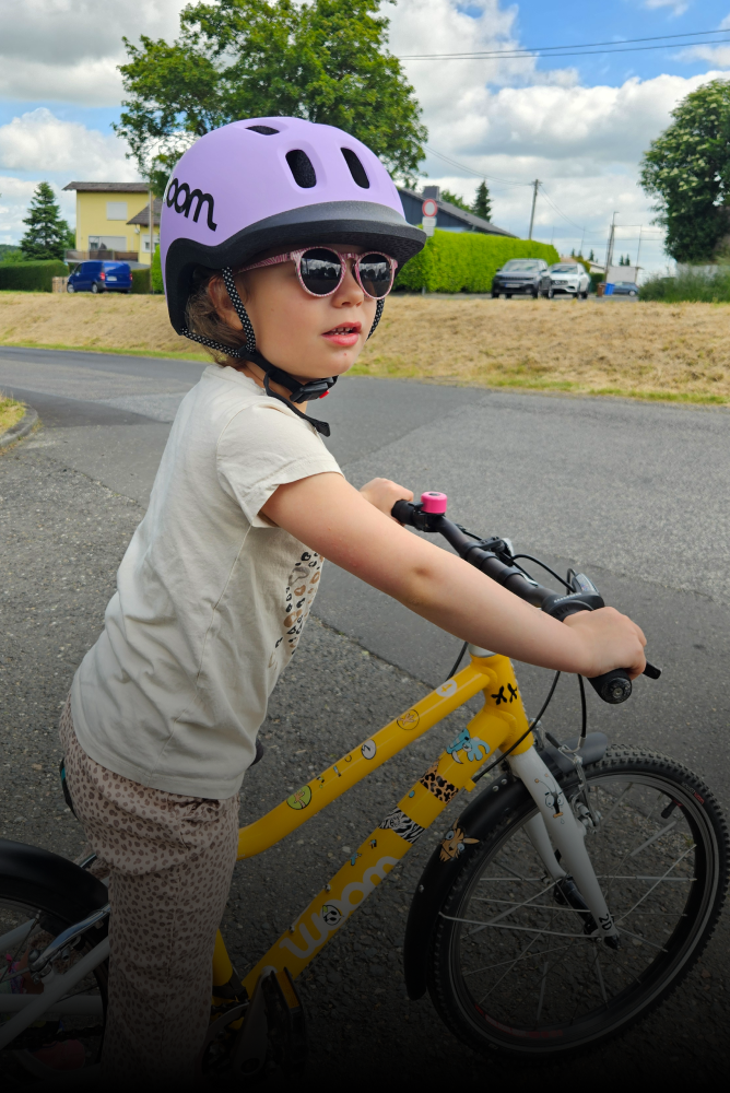 A child wearing a purple READY bike helmet is standing over a yellow woom bike with stickers all over the frame.