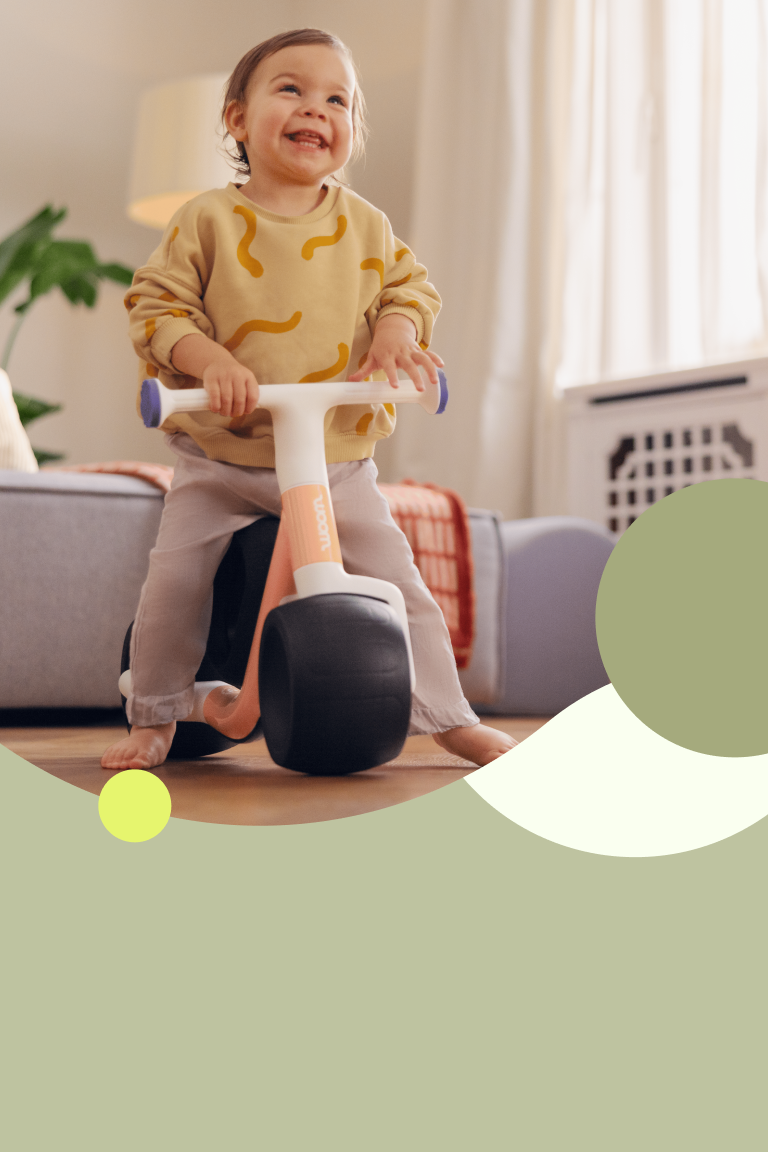 Breed grijnzende peuter op woom WOW loopfiets in pop peach.