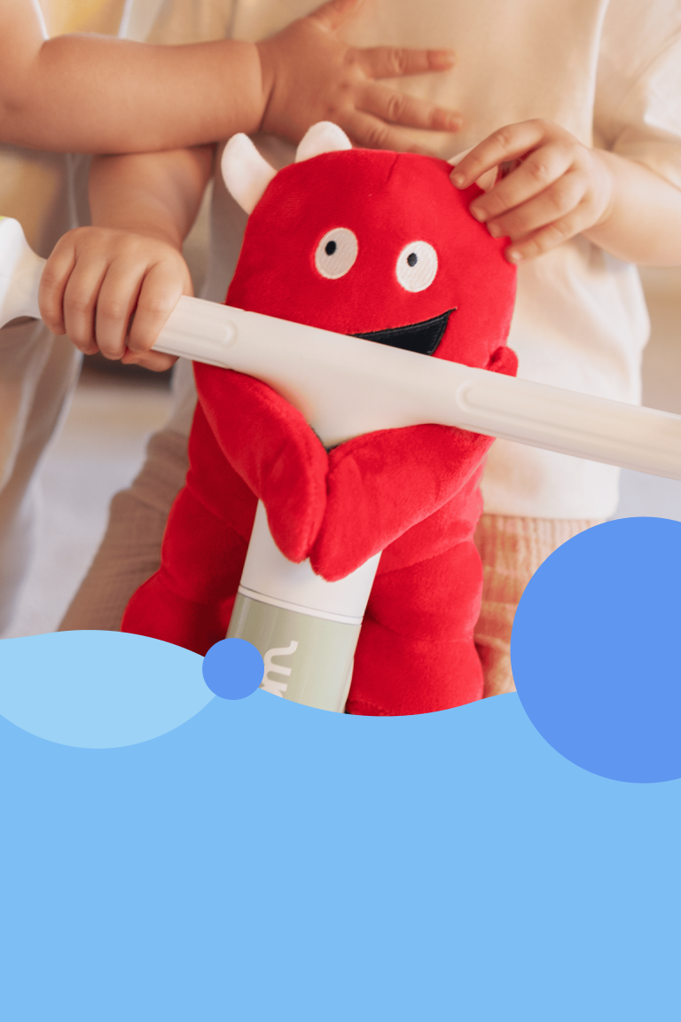The plush mascot Buddy is attached to the handlebars of the woom WOW balance bike. One toddler is sitting on the bike, another is standing next to him.