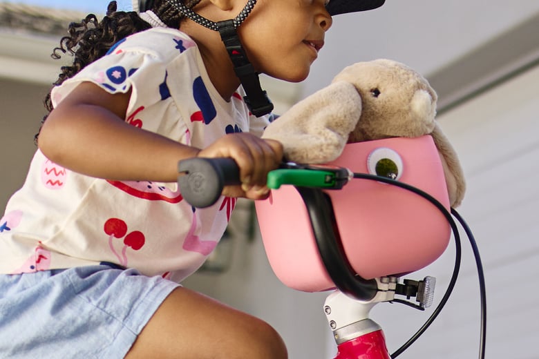 A young girl wearing a woom READY helmet rides a woom red GO bike with attached marshmallow pink POP basket holding a stuffed rabbit toy.