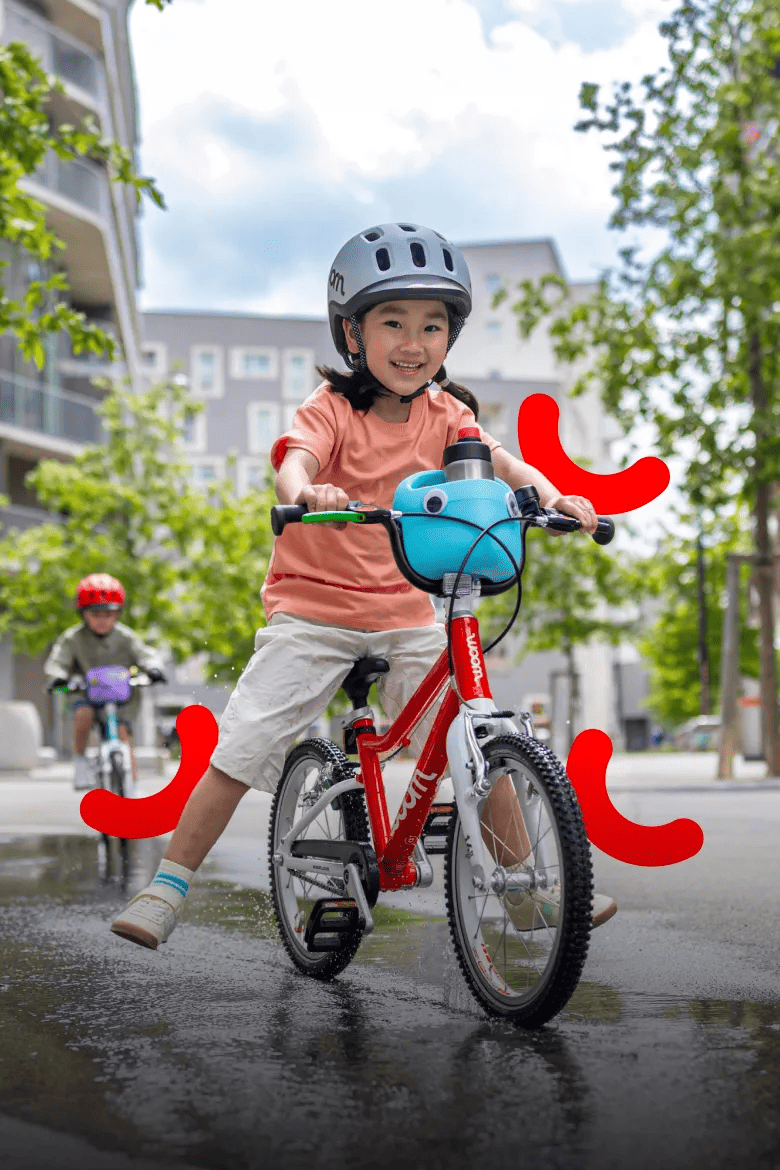 Kind mit woom READY Kinderhelm fährt mit einem roten woom GO 2 durch eine Pfütze.