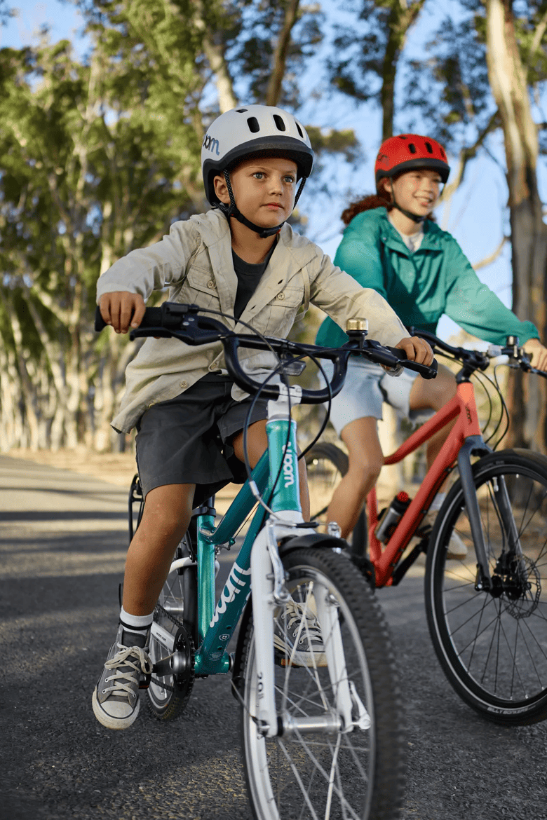 Zwei Kinder mit woom READY Helmen fahren auf einem woom GO und einem woom EXPLORE Bike einen baumbestandenen Weg entlang.