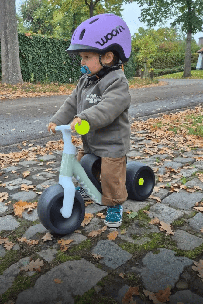 Małe dziecko w fioletowym kasku READY Kids jedzie na swoim zielonym rowerze samobalansującym woom WOW po brukowanym chodniku.