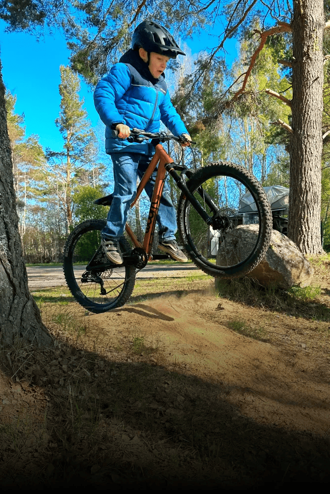 A boy is riding a copper-colored woom OFF AIR with the front wheel off the ground in a woodland setting.