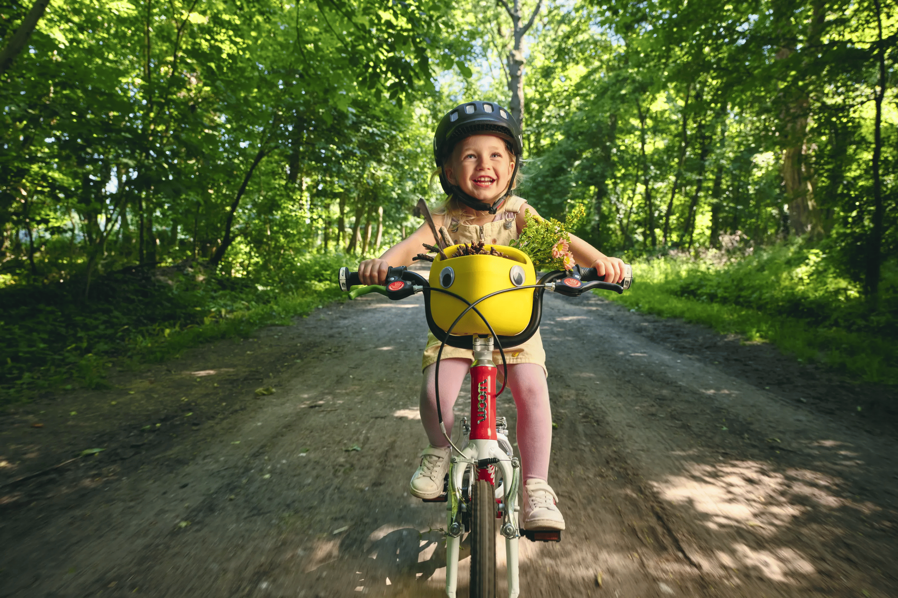 Ein kleines, lachendes Mädchen mit schwarzem woom Helm fährt auf einem rot-weißen woom bike auf einem schattigen Waldweg. Auf dem Lenker steckt ein gelber Fahrradkorb voll mit kleinen Schätzen aus der Natur.