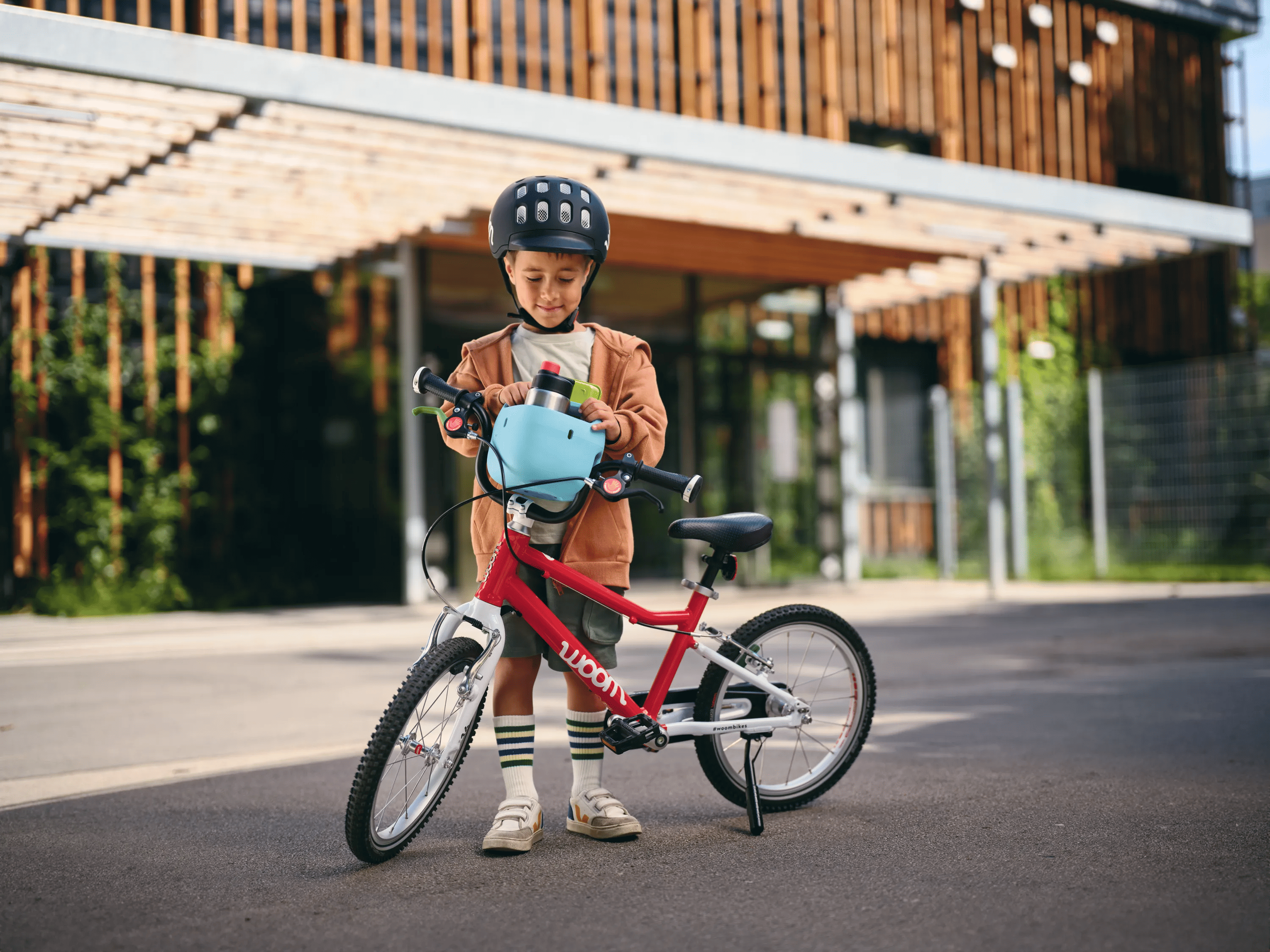 Ein kleiner Junge steckt den hellblauen woom Fahrradkorb fürs Kinderrad vorne auf den Lenker seines rot-weißen woom bikes.