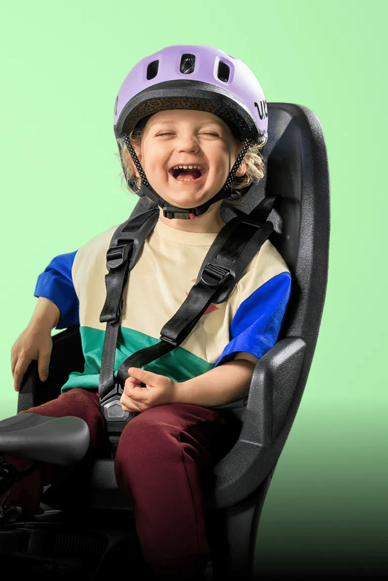 Strahlendes Kind mit READY Kinderhelm von woom in der Farbe Lila auf dem Kopf sitzt im Fahrradsitz vor grünem Hintergrund.