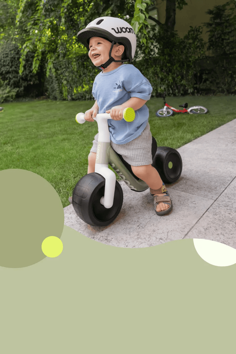 A happy child is riding into the shot on their woom WOW on the garden patio and wearing a READY Kids' Helmet.