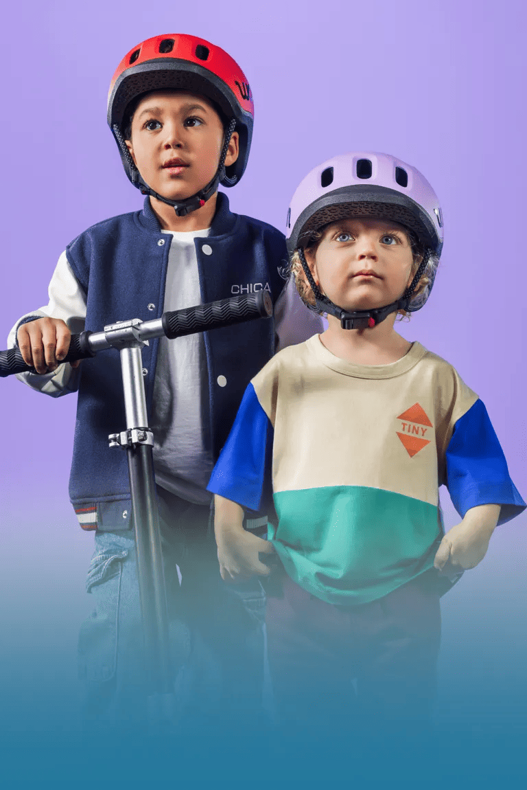 Zwei Kinder mit READY Kinderhelmen von woom auf den Köpfen und einem Scooter in der Hand blicken in die Ferne.
