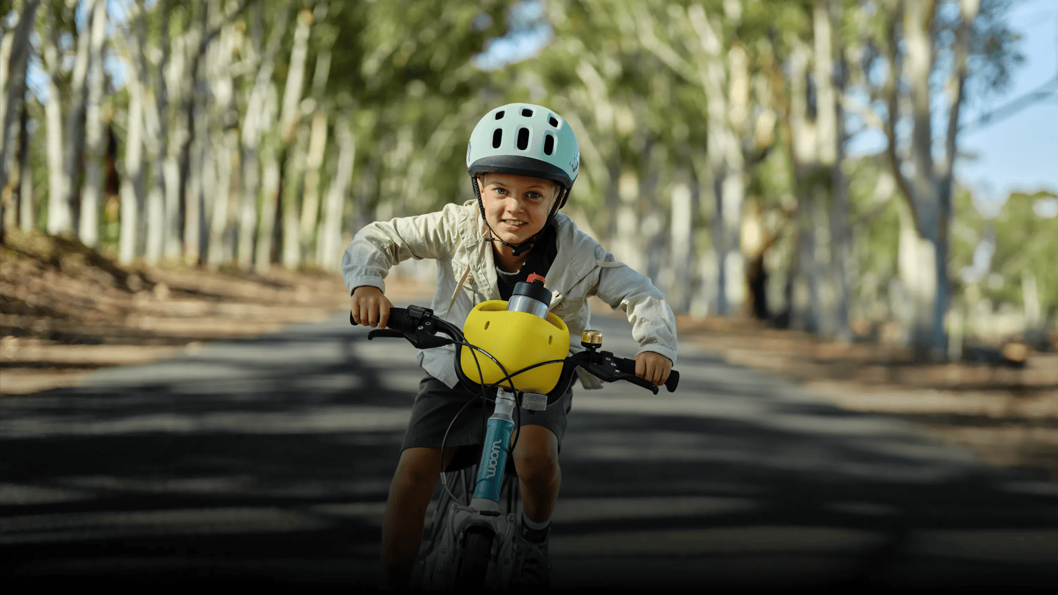 Kind mit READY Helm in Türkis auf dem Kopf fährt mit einem woom GO samt POP Fahrradkorb auf einer Straße im Grünen.
