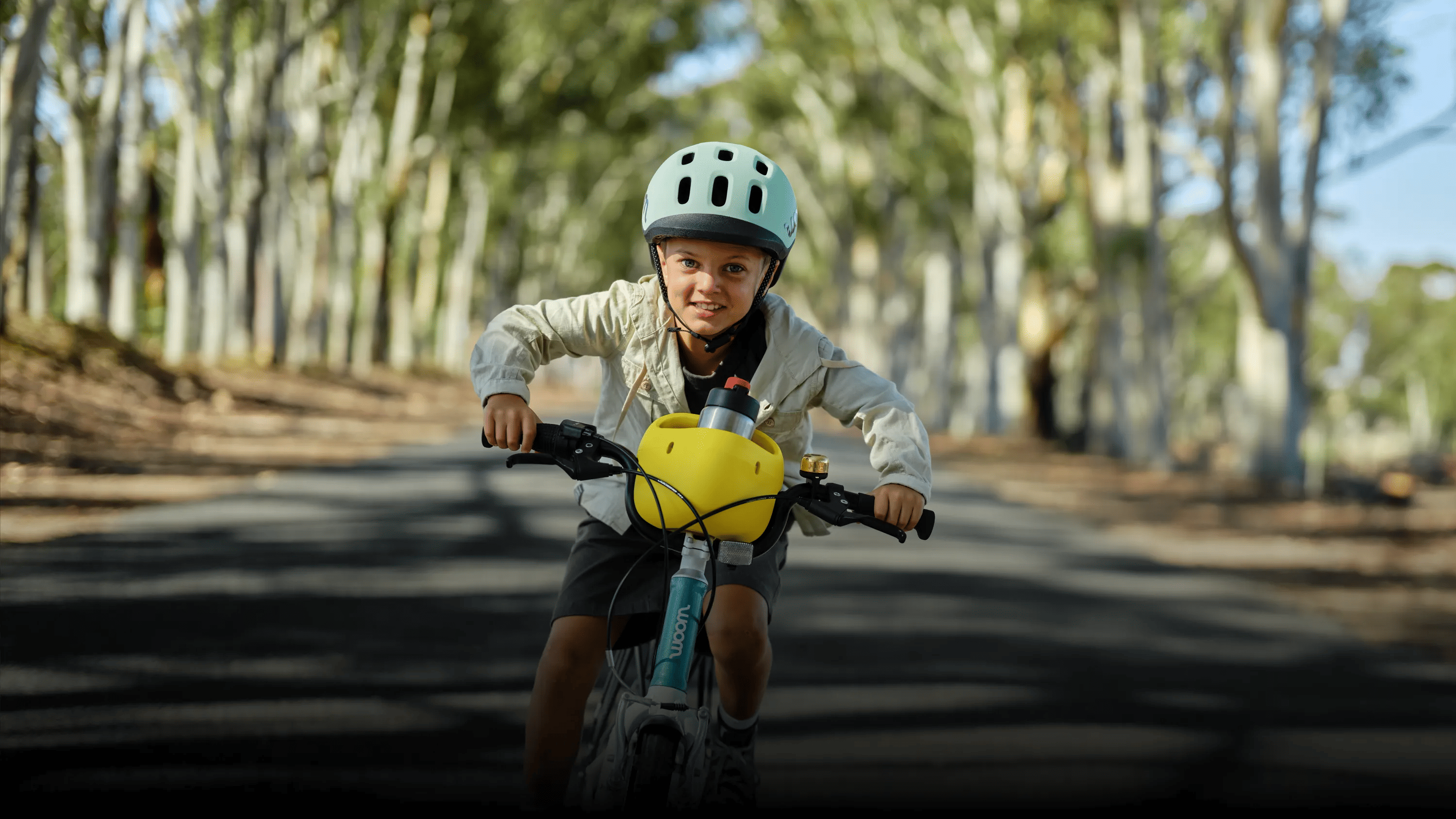 Kind mit READY Helm in Türkis auf dem Kopf fährt mit einem woom GO samt POP Fahrradkorb auf einer Straße im Grünen.