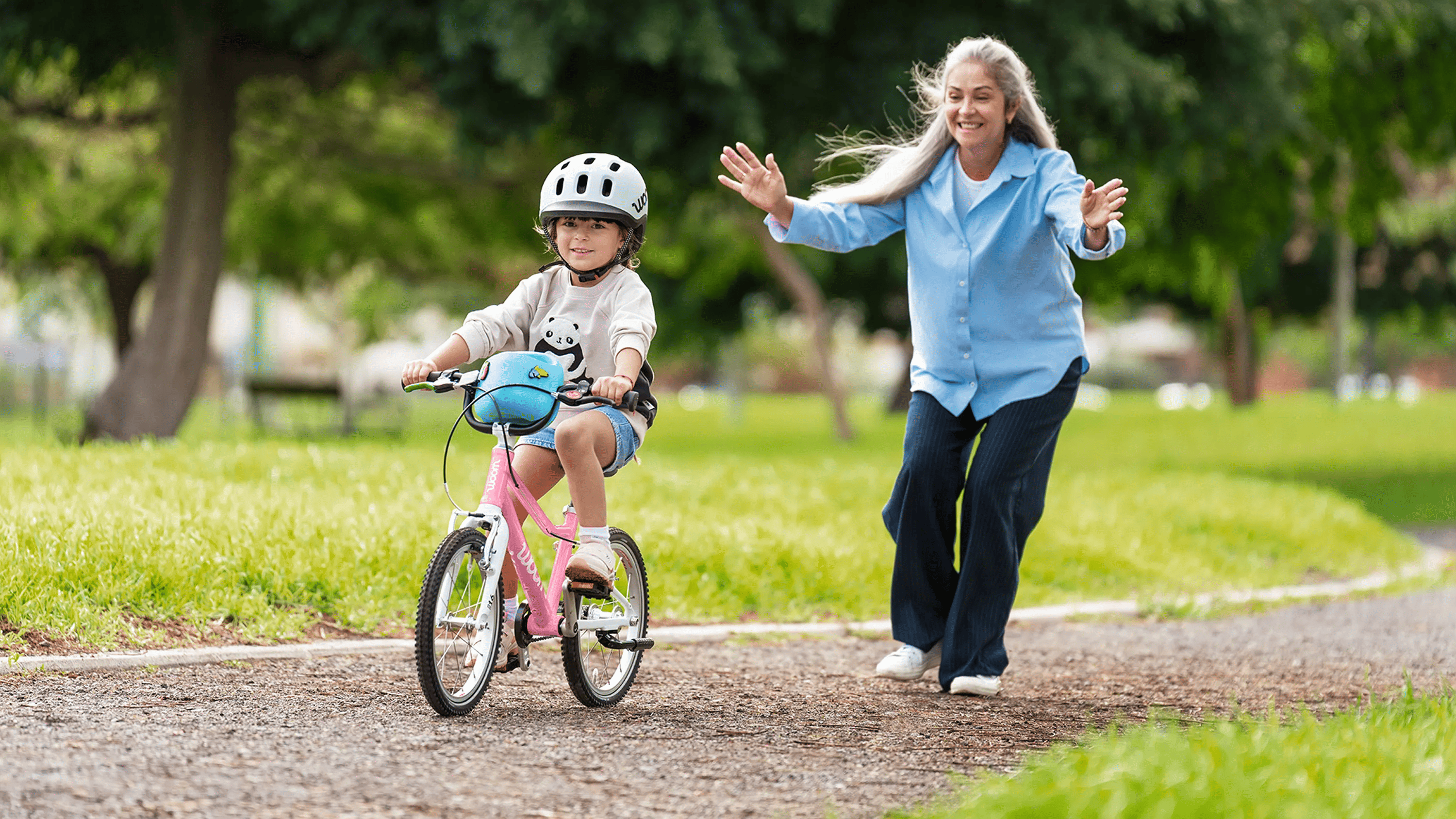 Kind met grijze READY-kinderhelm op woom GO-fiets in powder pink; vrouw juicht met open armen op een parkpaadje.