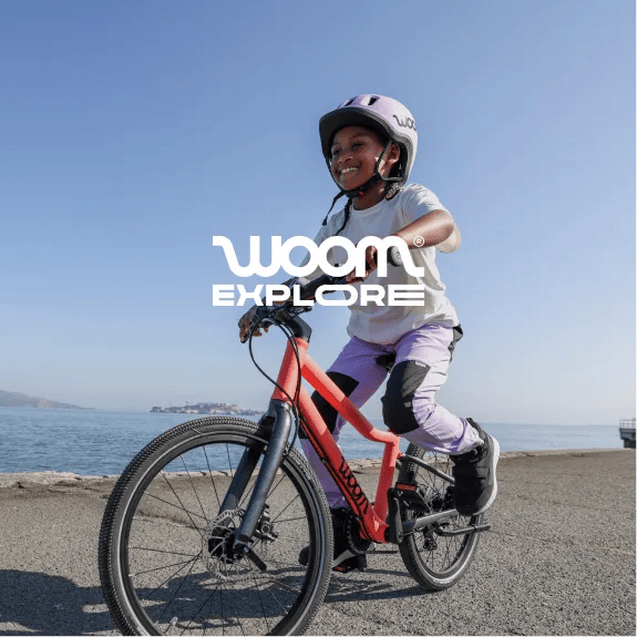 An image of a girl wearing a READY Helmet in lilac while riding her neon coral EXPLORE bike on a paved path next to a serene bay under a bright blue sky.