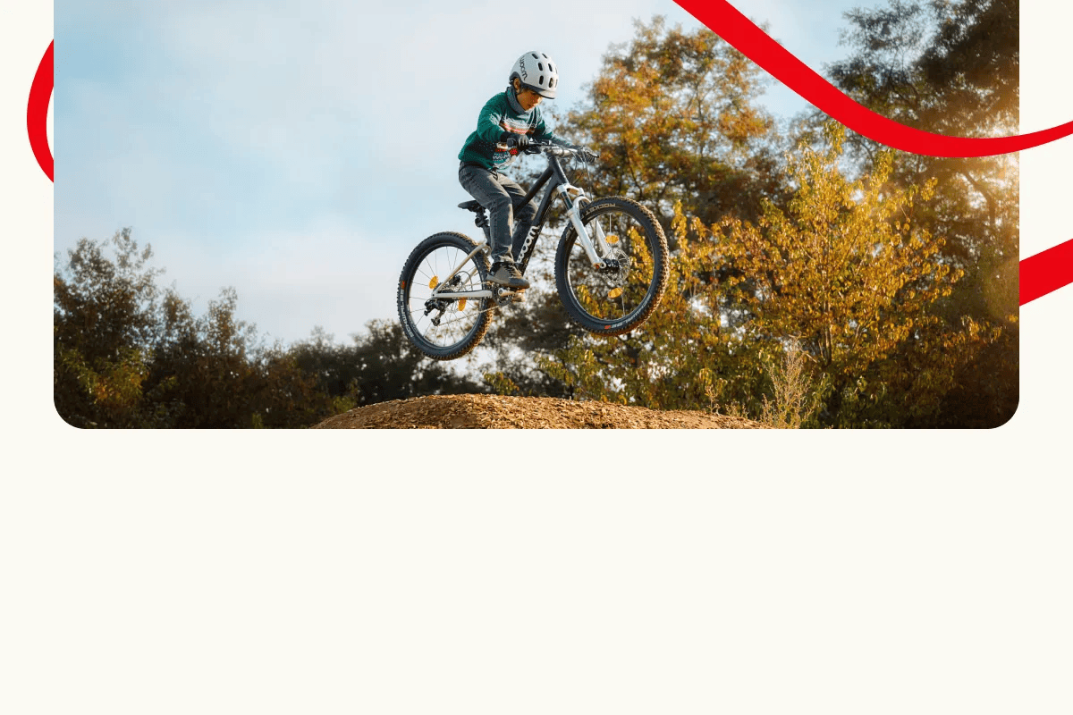Ein Kind auf einem Woom OFF Mountainbike springt hoch über eine Erdrampe, im Hintergrund sind Bäume und der Himmel zu sehen. Dekorative rote Linien umrahmen die Szene.