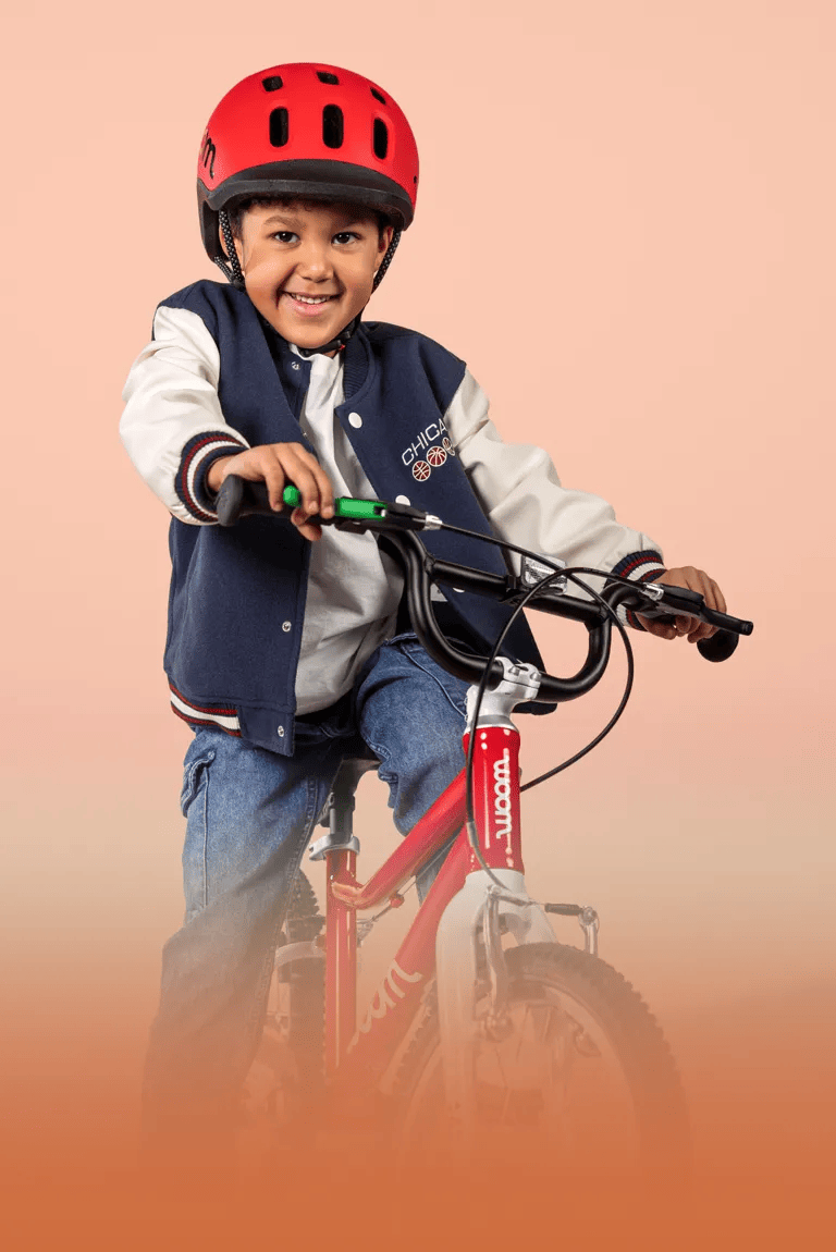 Auf dem Fahrrad woom GO in Rot sitzt ein frech lächelnder Junge mit rotem READY Kinderhelm auf dem Kopf.