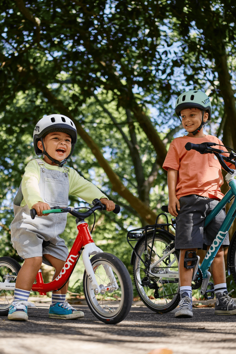 Zwei lächelnde Kinder mit woom READY Helmen auf ihren woom GO Bikes unter Bäumen.