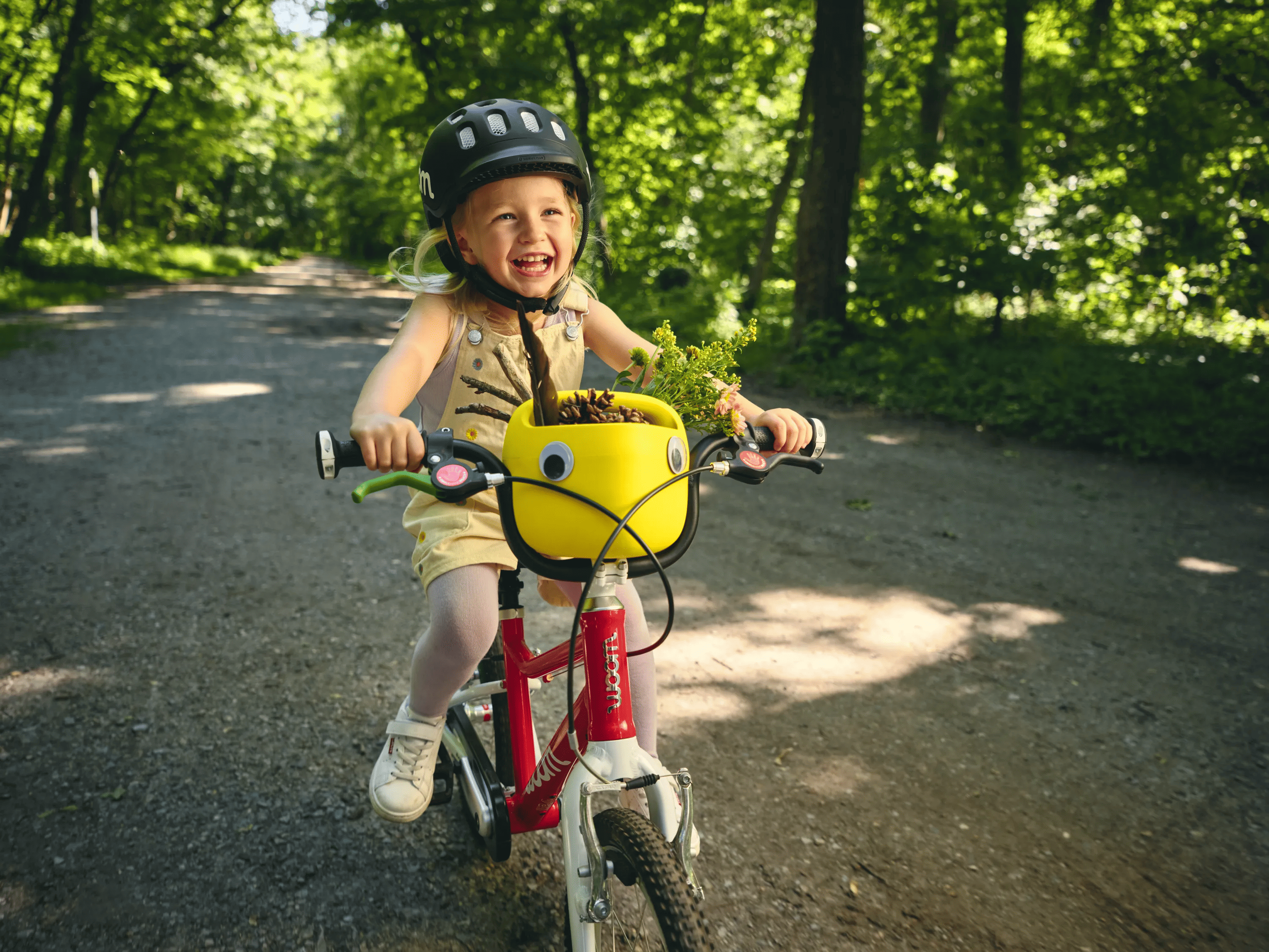 Ein kleines, lachendes Mädchen mit schwarzem woom Helm fährt auf einem rot-weißen woom bike auf einem schattigen Waldweg. Auf dem Lenker steckt ein gelber Fahrradkorb fürs Kinderrad.