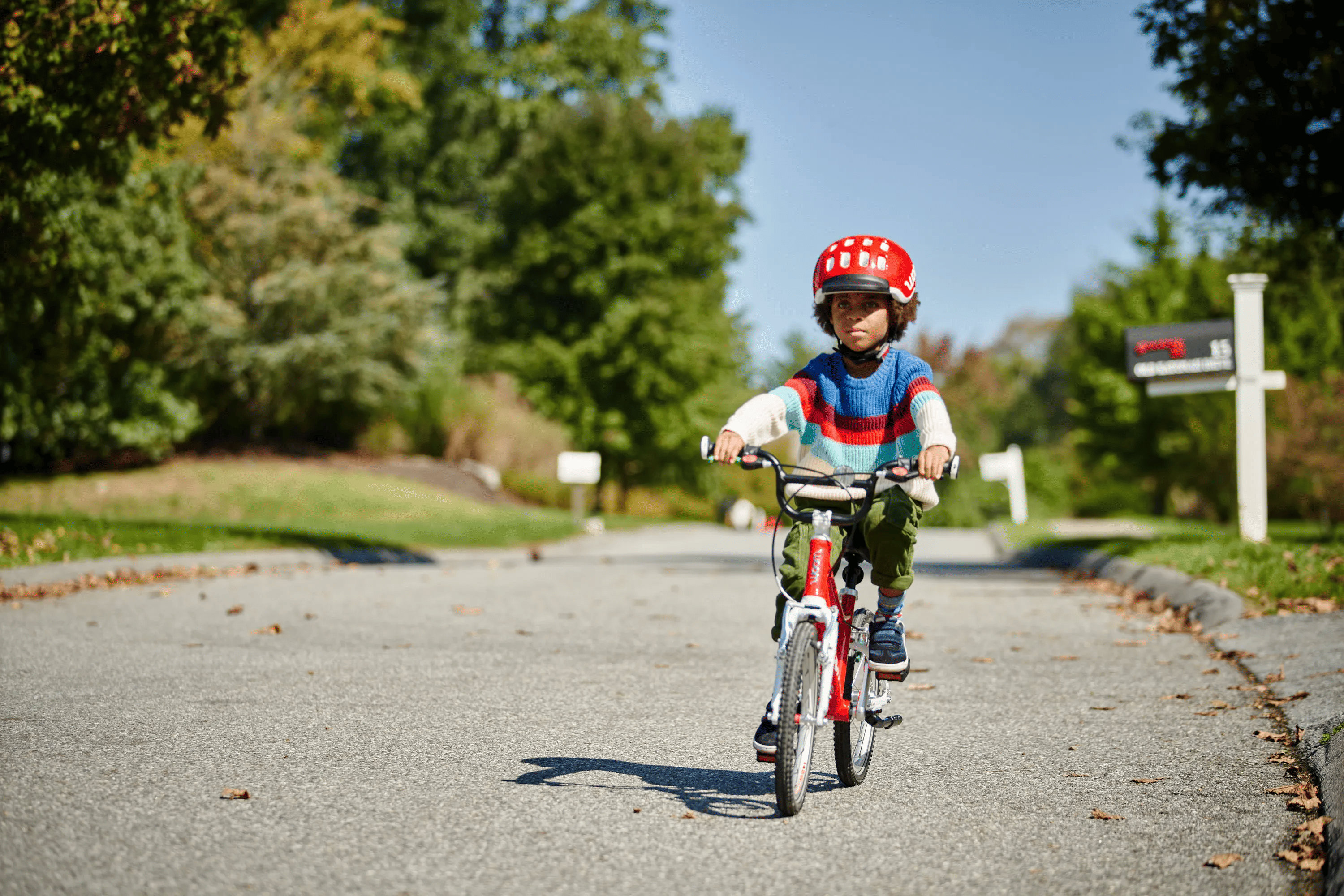 Child wearing a striped sweater and woom red KIDS' Helmet, riding a red woom ORIGINAL bike through a neighborhood.