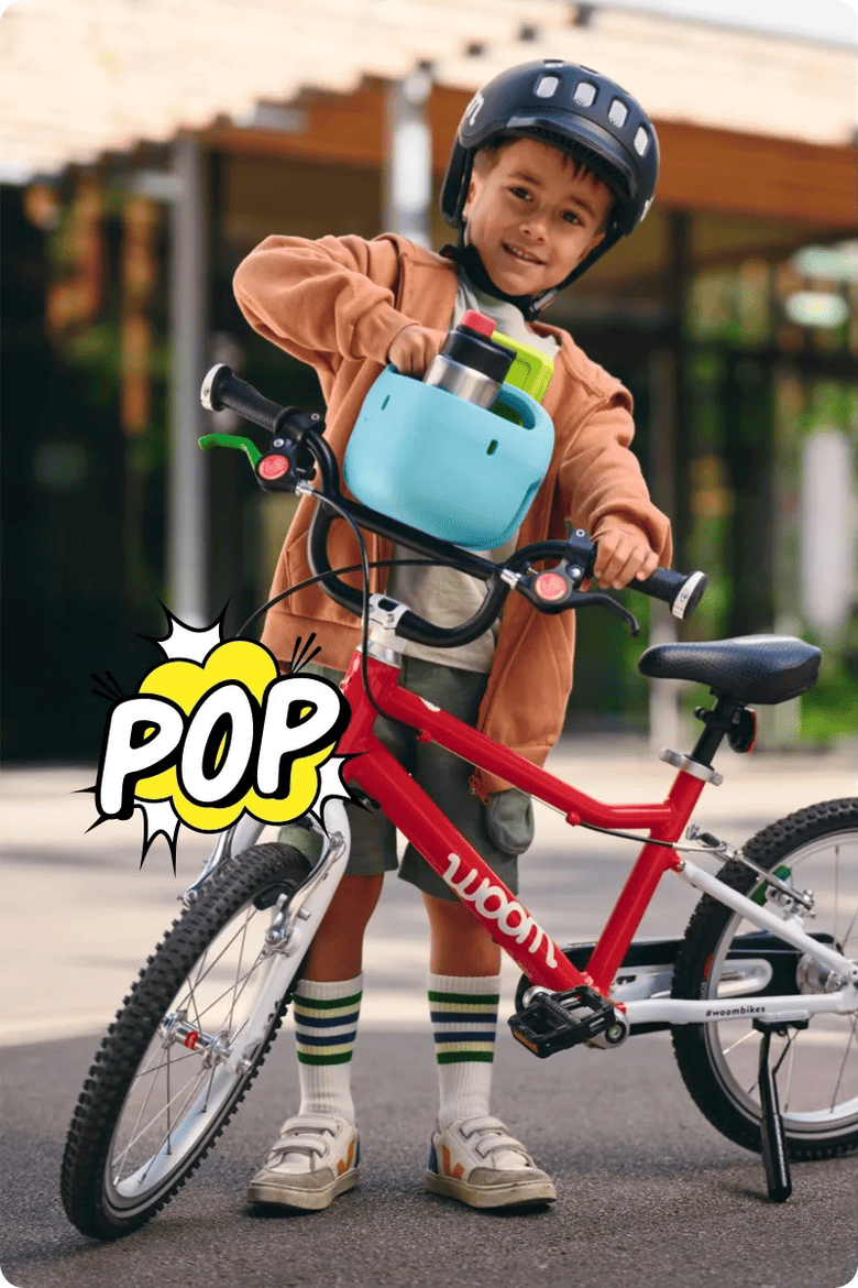 Ein kleiner Junge mit schwarzem Helm steckt einen hellblauen Fahrradkorb mit Trinkflasche vom Lenker seines rot-weißen woom bikes ab. Darunter ist eine zerplatzende Comic-Blase mit dem Schriftzug POP.