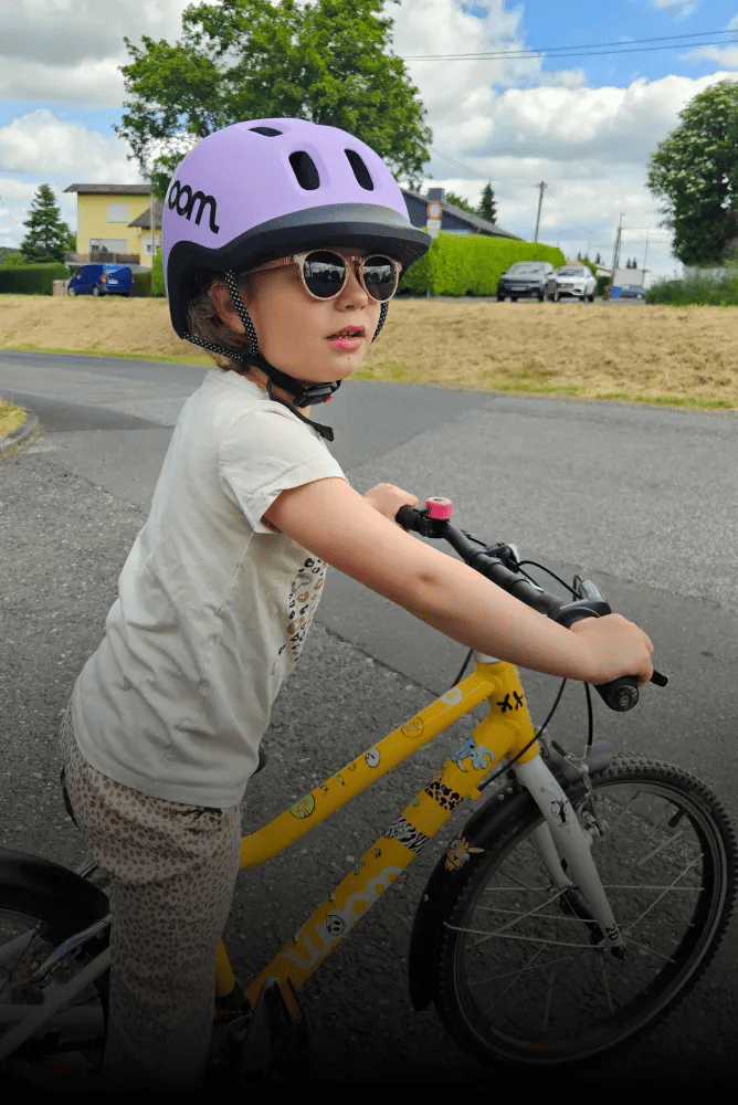Ein Kind mit lilafarbenem READY Radhelm steht auf einem gelben woom bike, dessen Rahmen mit Stickern beklebt ist.