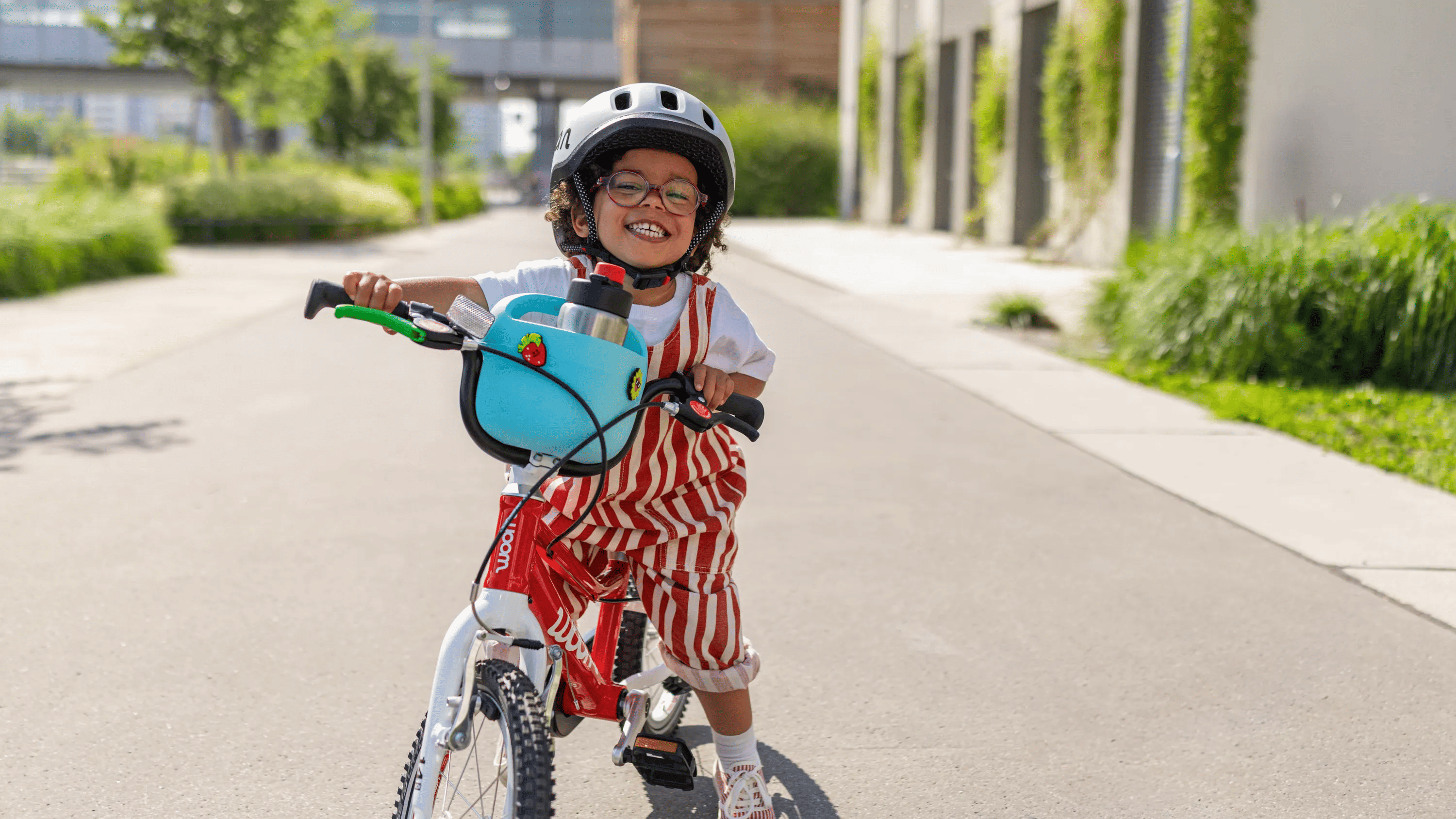 Smiling child riding a red woom GO bike with a blue woom POP bike basket and woom READY helmet on a sunny path.