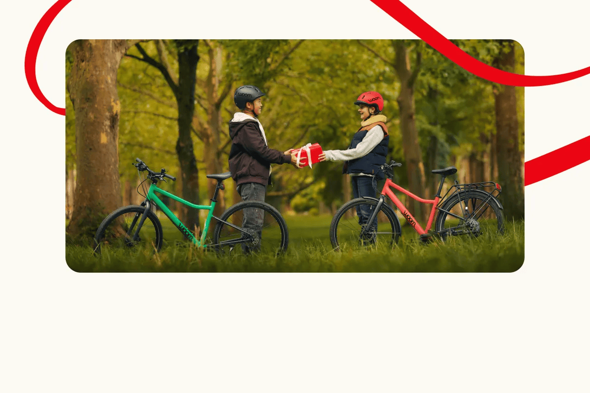 Two kids with woom EXPLORE bikes in a forest are exchanging a gift. Decorative red lines frame the scene.