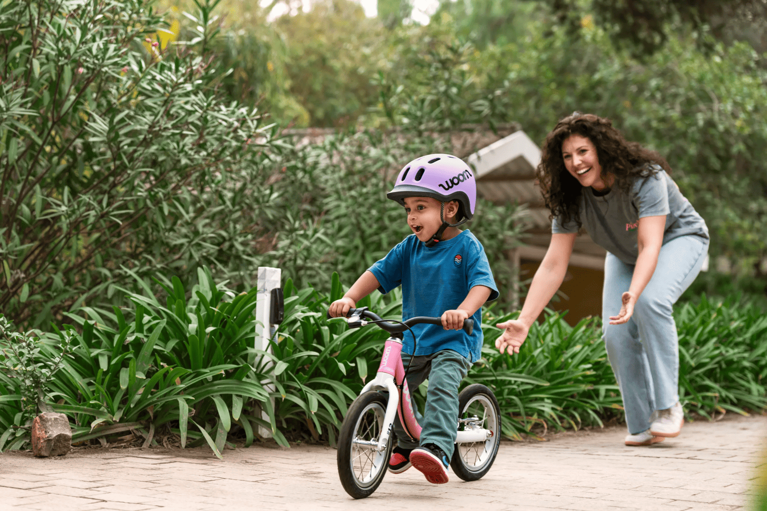 Child in a lilac woom READY helmet on a powder pink woom GO 1 bike, with a woman cheering on a garden path.