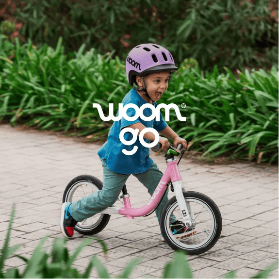 An image of a young boy grinning and wearing a READY Helmet in lilac while riding a powder pink woom GO 1 down a brick path.