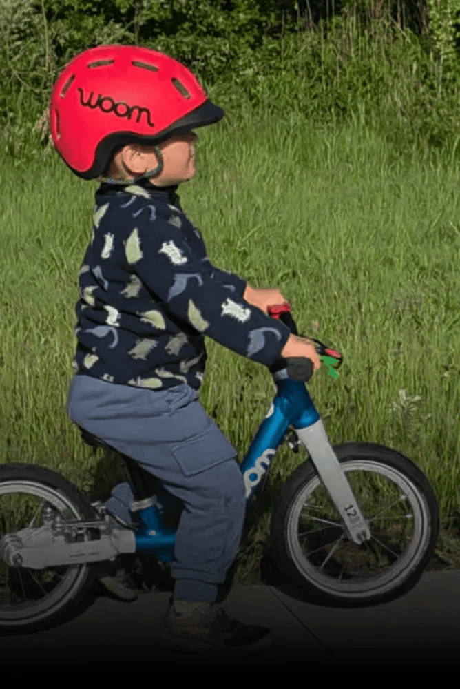Ein Kind mit rotem READY Radhelm rollt auf einem blauen Laufrad von woom, im Hintergrund ist eine Wiese.