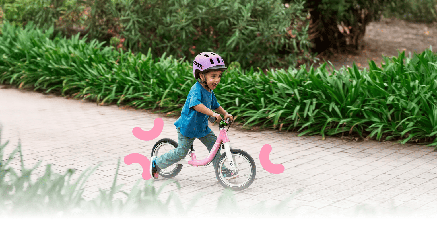 Child with a purple READY helmet rides a pink woom GO bike along a paved path. Green, blooming bushes in the background.