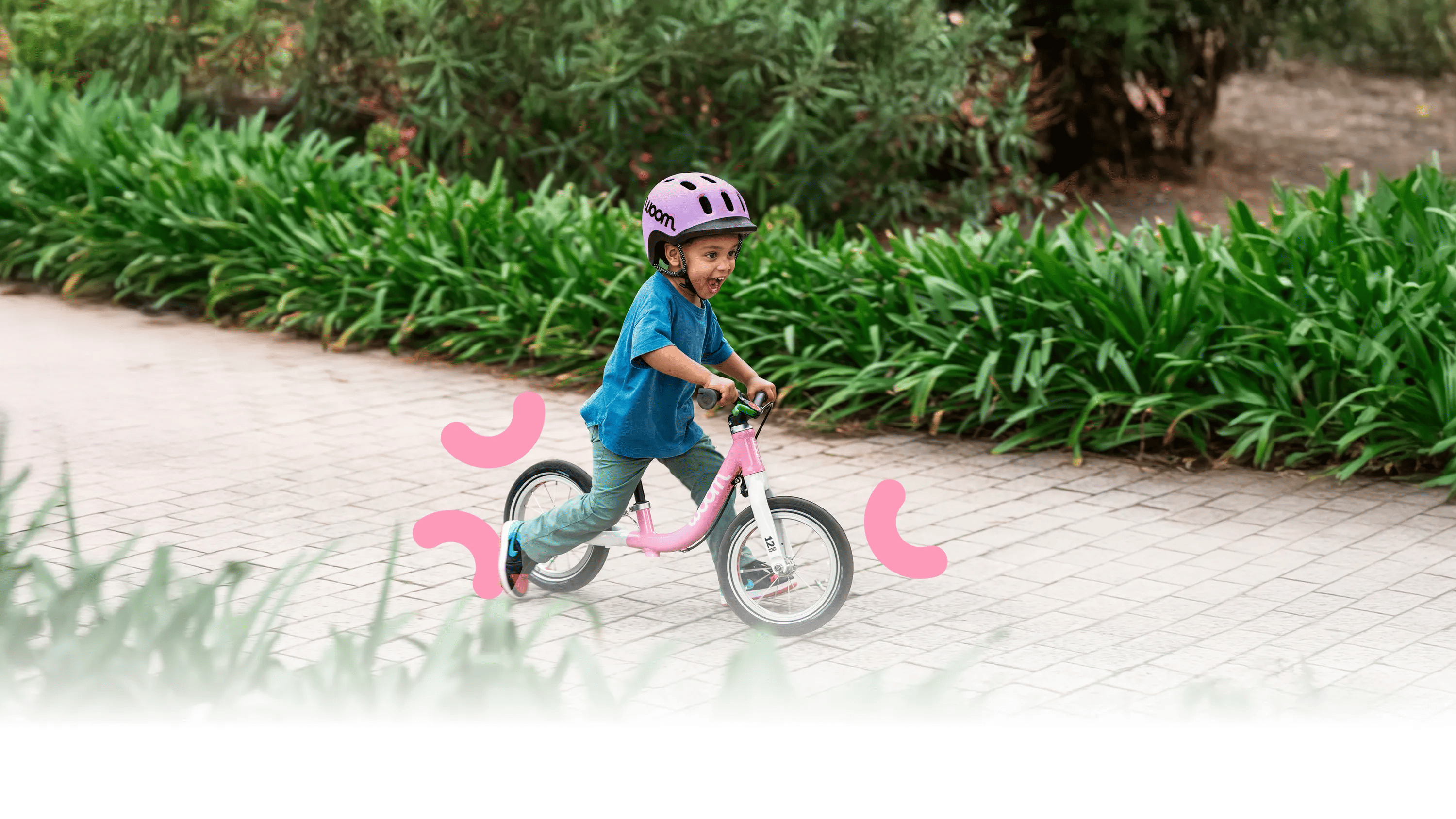 Child with a purple READY helmet rides a pink woom GO bike along a paved path. Green, blooming bushes in the background.