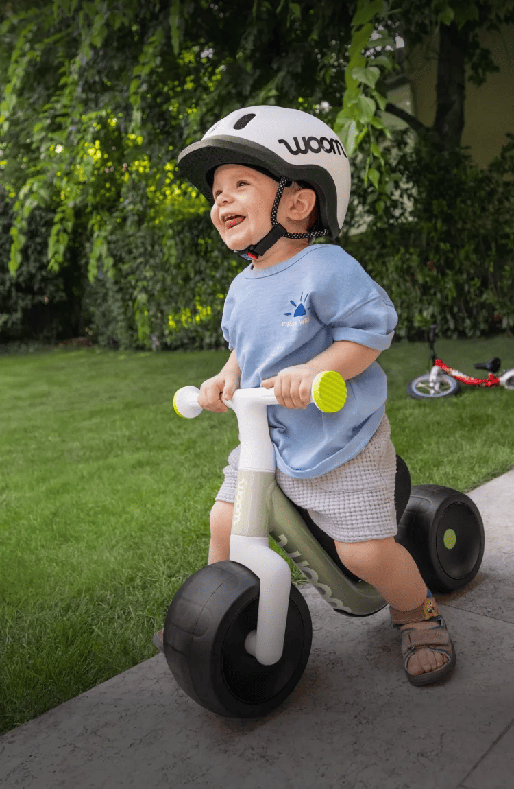 Smiling child with helmet on a woom balance bike in a garden. Ad banner for woom WOW and woom GO models.