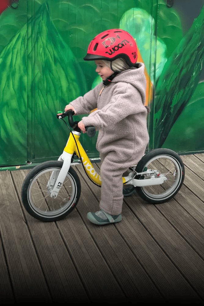 Auf einem gelben woom Laufrad in 12 Zoll rollt ein Kleinkind mit rotem READY Radhelm und Wollwalkanzug.