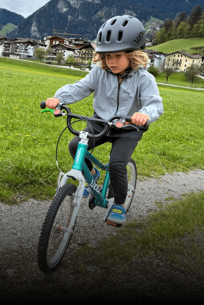 Kind mit schwarzem READY Helm fährt mit einem grünen woom GO Fahrrad auf einem Schottertrail durch eine Wiese in den Bergen.